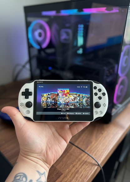 Person holding a PlayStation Vita with a gaming screen in front of a computer monitor. White Sony PS Vita handheld console with custom firmware, pre-installed games, emulators, and 128GB SD card.